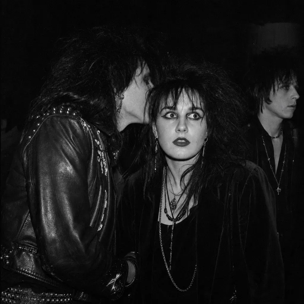 Black-and-white flash photograph in a dark club: a goth couple pressed close, one whispering into the other's ear, a third figure watching from the shadow.