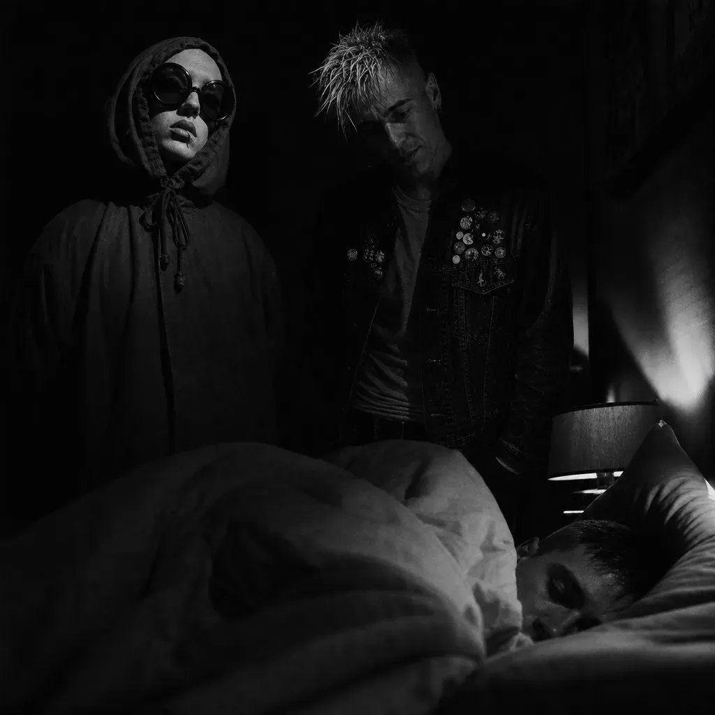 Two figures — a hooded girl in sunglasses and a pin-jacketed young man — stand over a stranger's bed at night, leaning slightly toward the sleeper.