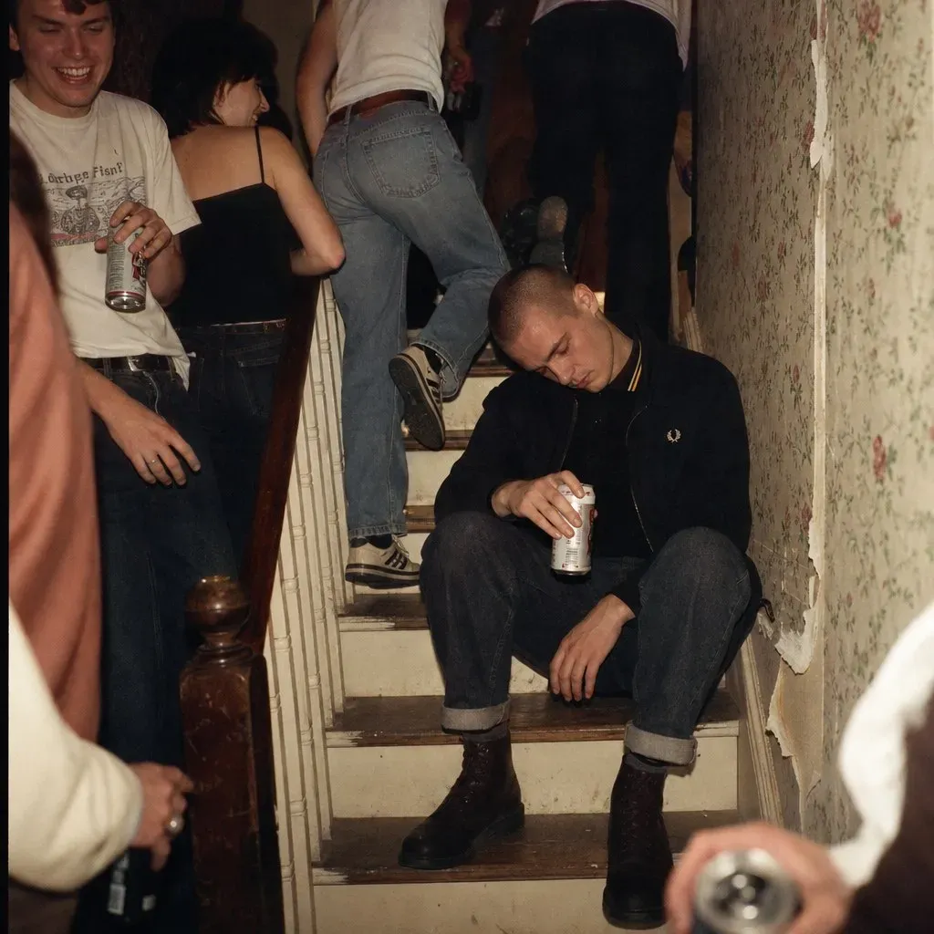 A houseparty stairwell: someone stepping over a young man asleep sitting up, beer can still in hand, wallpaper peeling behind him.