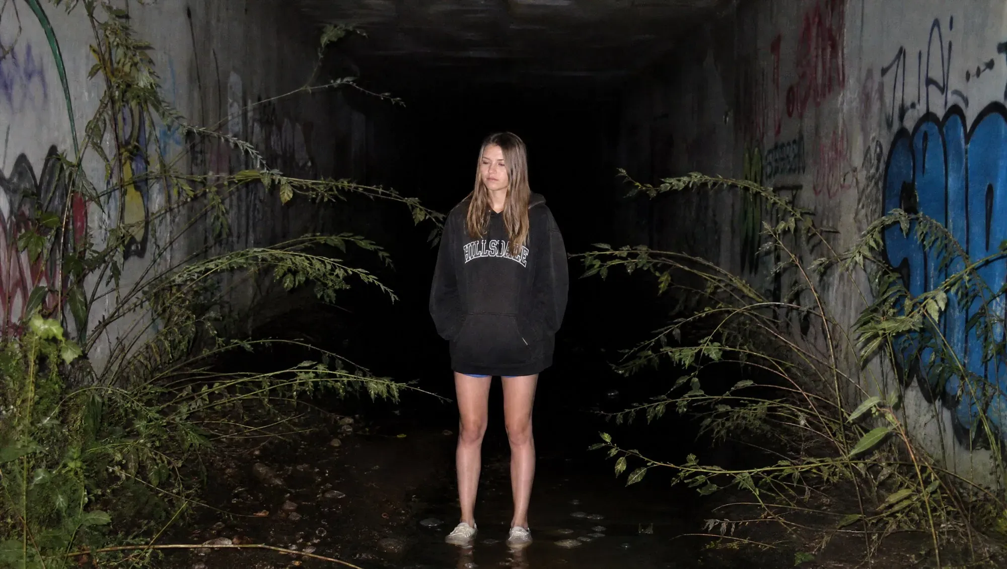Direct-flash night photograph. A teenage girl stands ankle-deep in water at the mouth of a graffitied, overgrown concrete tunnel, arms at her sides.