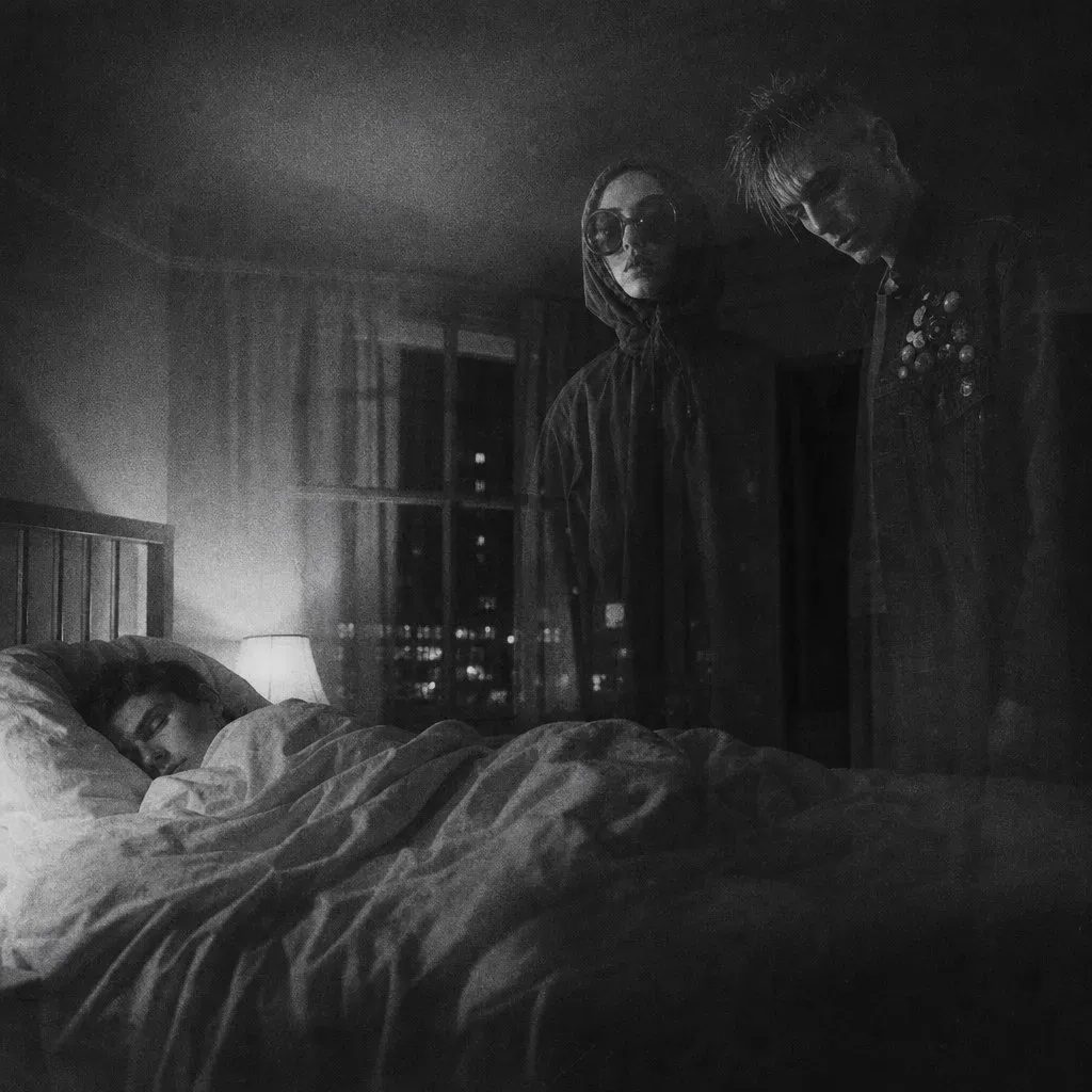 Two figures at the foot of a bed in a dark flat, a sleeper curled under the blanket, the city lit in the window behind them.