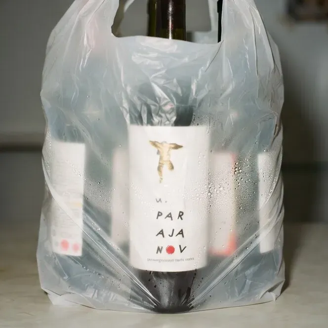 A plastic carrier bag on a kitchen counter, sweating with condensation, full of bottles of "M. Parajanov" semi-sweet pomegranate wine — the gold label showing a small spread-armed figure — a receipt tucked in among them.
