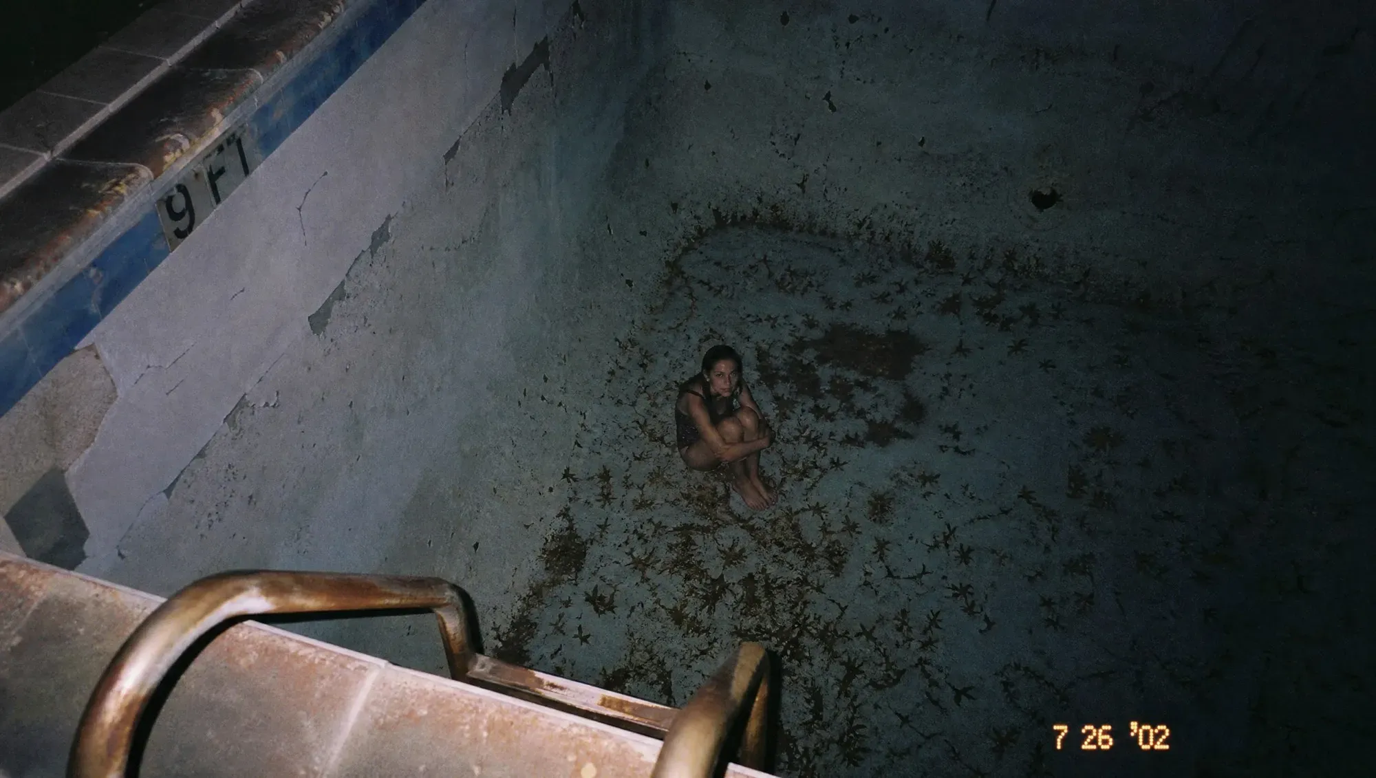 Disposable-camera photo, date-stamped 7 26 '02. Shot from the edge of a drained swimming pool at night; a girl sits alone in the deep end among dead leaves.