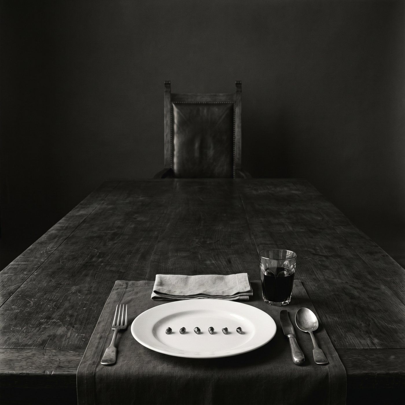 A long dark table set for one — a single plate, cutlery, a glass of near-black wine — in a near-black room.