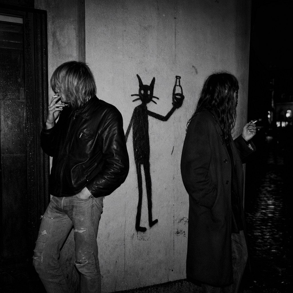 Black-and-white flash photo: two punks smoke against a pale wall, and painted between them a thin black cat figure raises a bottle.