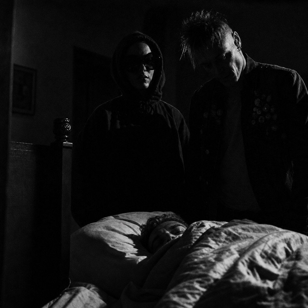 Two figures standing calm at a sleeper's bedside in the dark, patient as furniture.