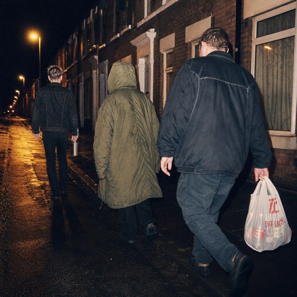Three figures walking away down a terraced street at night, one in a long parka, one carrying a plastic bag — the walk home at the hour nobody owns.